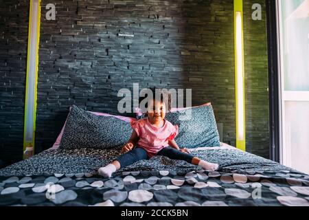 Jeune fille souriante avec les jambes écartées assis sur le lit contre mur à la maison Banque D'Images