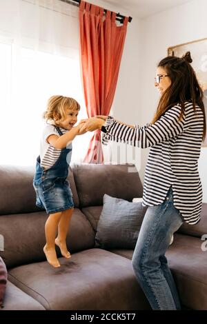 Une mère heureuse tenant les mains de sa fille joueuse sautant sur son canapé accueil Banque D'Images