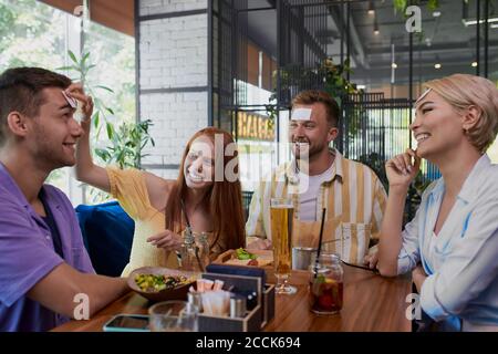 petit autocollant sur le front. amis s'amusant dans le restaurant, jeunes caucasiens jouant un jeu hedbanz, rire Banque D'Images