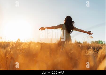 Jeune femme aux bras tendus debout au milieu d'une ferme de blé contre ciel dégagé au coucher du soleil