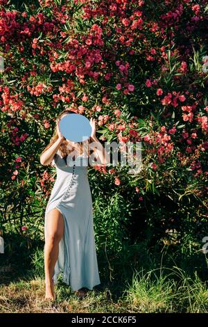 Jeune femme portant une robe tenant un miroir debout près des plantes dans le parc le jour ensoleillé Banque D'Images