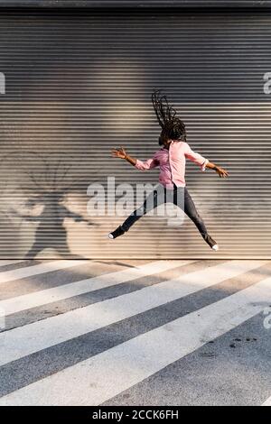 Jeune homme afro avec des dreadlocks sautant dans la rue contre le mur Banque D'Images