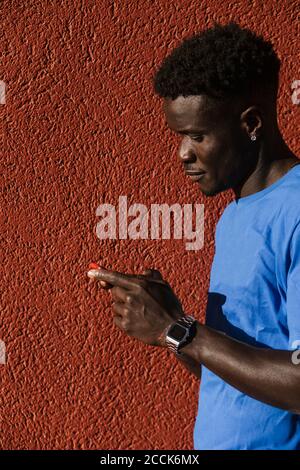 Gros plan d'un jeune homme utilisant un smartphone contre un mur rouge en ville pendant la journée ensoleillée Banque D'Images