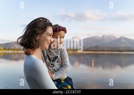 Mère souriante portant une jolie fille tout en se tenant contre le lac à coucher de soleil Banque D'Images