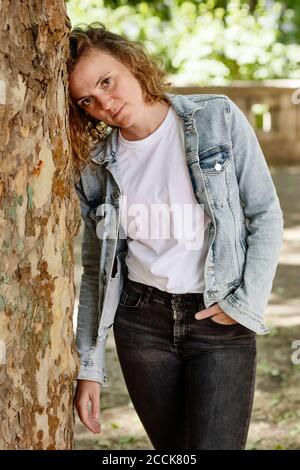 Femme adulte de taille moyenne portant une veste en denim penchée sur le tronc d'arbre dans le parc Banque D'Images