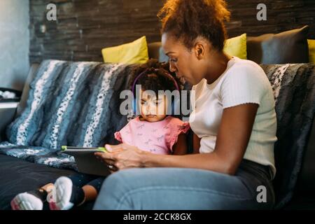Mère montrant une tablette numérique à fille portant un casque pendant qu'elle est assise sur le canapé à la maison Banque D'Images
