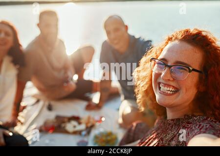 Bonne femme à tête rouge ayant pique-nique avec des amis sur la jetée à un lac au coucher du soleil Banque D'Images