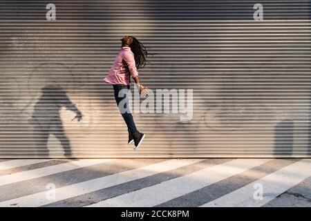 Jeune homme insouciant avec des dreadlocks sautant dans la rue contre le mur Banque D'Images