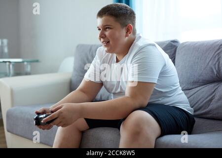 fat caucasien adolescent garçon aime les jeux vidéo, le garçon en surpoids est assis sur le canapé tenant la console dans les mains, concentré sur le jeu, regarder l'écran Banque D'Images