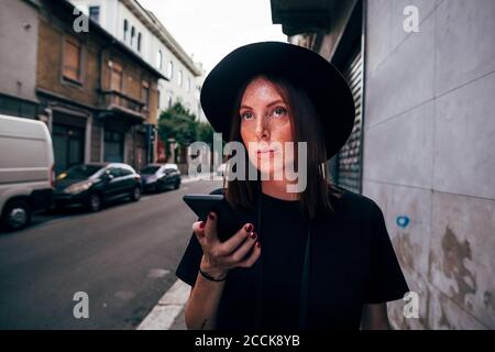 Jeune femme portant un chapeau tenant un smartphone tout en regardant loin en ville Banque D'Images
