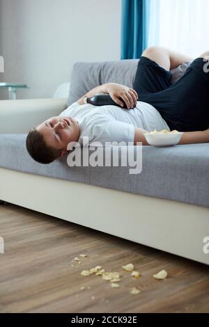 un garçon en surpoids dormait sur un canapé dans la salle de séjour, un jeune garçon de race blanche s'endormit pendant qu'il regardait la télévision et mangeait des chips Banque D'Images