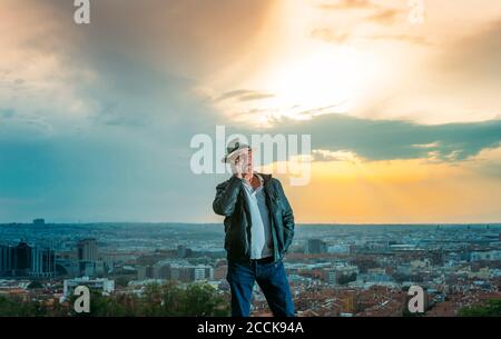 Homme senior parlant sur téléphone mobile avec la ville en arrière-plan au coucher du soleil Banque D'Images