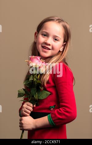 Jeune jolie fille de 8 ans vêtue d'une robe rouge tenant des fleurs isolées sur fond de couleur orange Banque D'Images