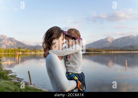 Mère portant fille joyeuse en se tenant au bord du lac contre le ciel au coucher du soleil Banque D'Images