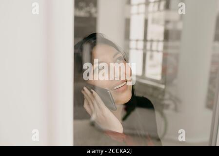 Portrait d'une femme souriante utilisant un smartphone derrière la fenêtre Banque D'Images