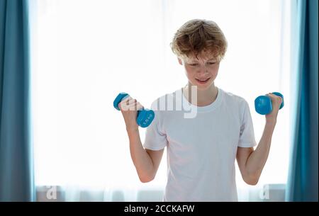 jeune caucasien garçon adolescent fort s'exerçant à la maison, tenir des haltères dans les mains, pompant les muscles de bras. concept de sport Banque D'Images