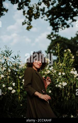 Femme adulte de taille moyenne avec cheveux bouclés portant un manteau debout contre plantes dans le parc Banque D'Images