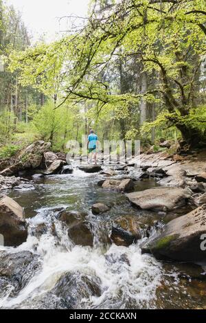 Homme senior debout sur le rocher au milieu de la rivière Rur Au printemps, vous traversiez High Fens - Eifel nature Park Banque D'Images