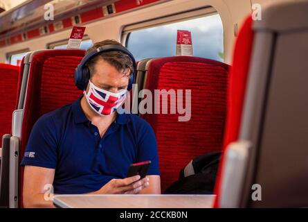 Un homme voyage dans un train portant un masque Union Jack et un polo bleu sur fond de sièges rouges. Banque D'Images
