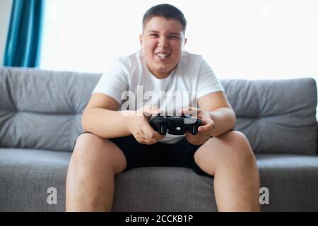 fat caucasien adolescent garçon aime les jeux vidéo, le garçon en surpoids est assis sur le canapé tenant la console dans les mains, concentré sur le jeu, regarder l'écran Banque D'Images