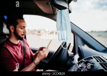 Gros plan du masque suspendu sur le rétroviseur pendant que l'homme utilisation du téléphone en voiture Banque D'Images