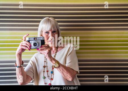 Femme âgée élégante qui photographie avec un appareil photo tout en se tenant contre le mur à la maison Banque D'Images