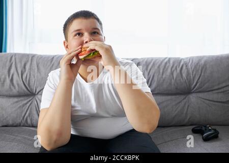 portrait d'un gros garçon mangeant un sandwich et regardant la télévision à la maison. dans la salle de séjour Banque D'Images