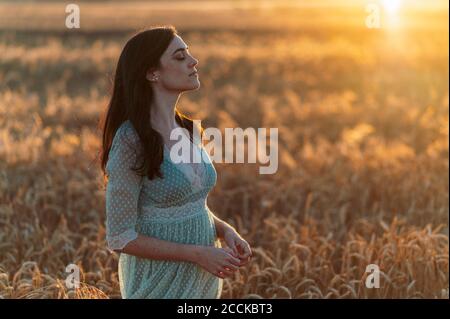 Jeune femme avec les yeux fermés debout au milieu des récoltes de blé dedans ferme au coucher du soleil Banque D'Images