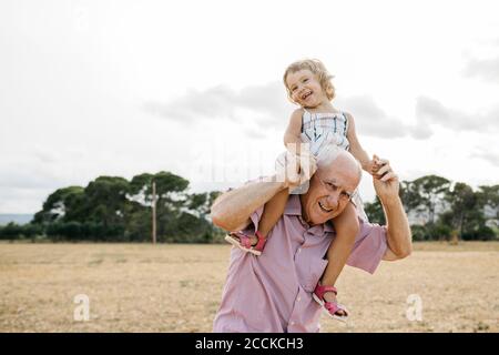 Grand-père heureux portant la petite-fille sur les épaules tout en étant debout contre le ciel Banque D'Images