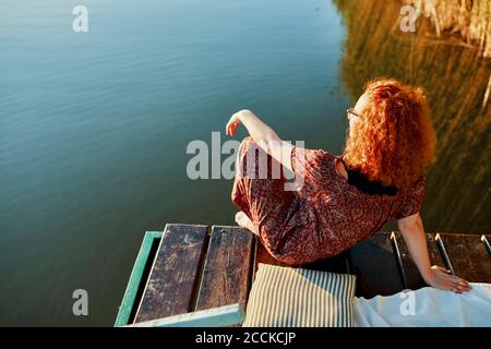 Jeune femme à tête rousse assise sur une jetée à un lac à coucher de soleil Banque D'Images