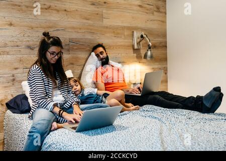 Fille couchée par des parents travaillant sur des ordinateurs portables sur le lit à accueil Banque D'Images