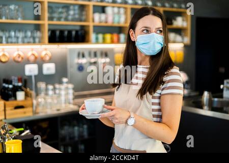 Jeune propriétaire attentif portant un masque tout en tenant une tasse de café et soucoupe dans le café Banque D'Images