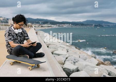 Jeune homme utilisant un smartphone assis avec un skateboard mur de soutènement par mer Banque D'Images