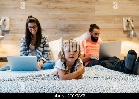 Jolie fille couchée sur le lit pendant que la mère travaille sur un ordinateur portable et le père s'est endormi dans la chambre à la maison Banque D'Images