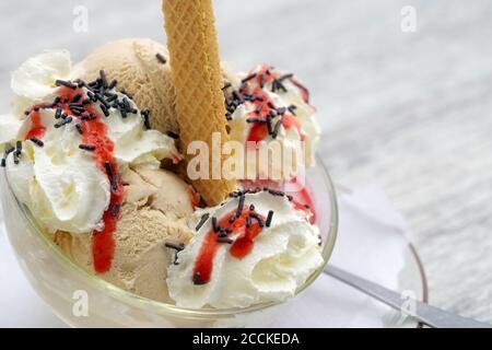 Détail d'une sundae avec des boules de crème glacée, de la crème fouettée, de la sauce aux fruits rouges et des saupoudrés de chocolat dans un bol en verre sur une table grise, espace de copie, gros plan Banque D'Images