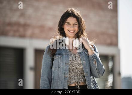 Jeune femme souriante avec des écouteurs debout en ville sous le soleil jour Banque D'Images