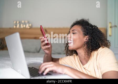 Jeune femme joyeuse utilisant un smartphone et un ordinateur portable sur le lit à la maison Banque D'Images