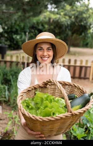 Jeune femme souriante tenant un panier en osier avec des légumes dans la cour pendant le couvre-feu Banque D'Images