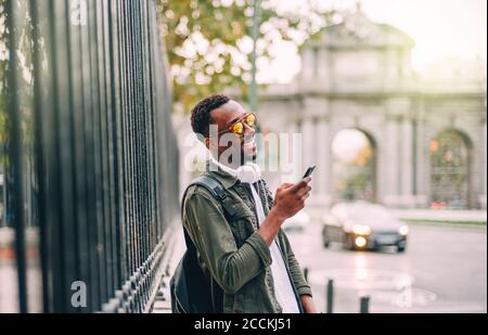 Homme souriant portant des lunettes de soleil à l'aide d'un smartphone lorsqu'il est debout ville Banque D'Images