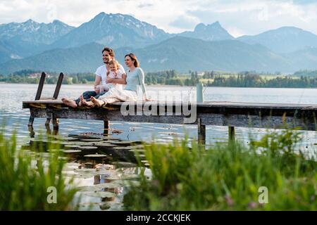 Une famille heureuse qui regarde la vue tout en étant assise sur la jetée lac contre les montagnes Banque D'Images