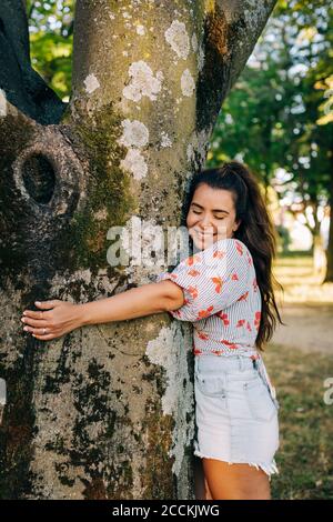 Jeune femme souriante avec les yeux fermés embrassant le tronc d'arbre pendant en position de stationnement