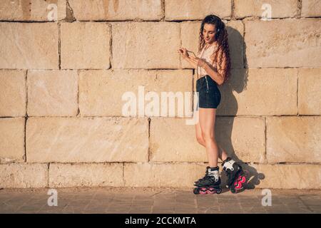 Jeune femme patineuse en ligne en utilisant un smartphone appuyé sur un mur Banque D'Images