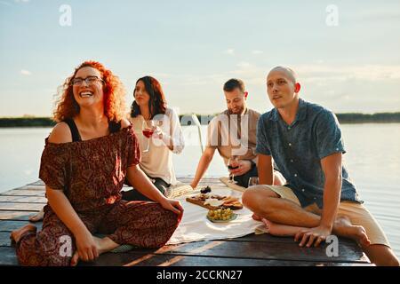 Des amis heureux ayant pique-nique sur la jetée à un lac à coucher de soleil Banque D'Images