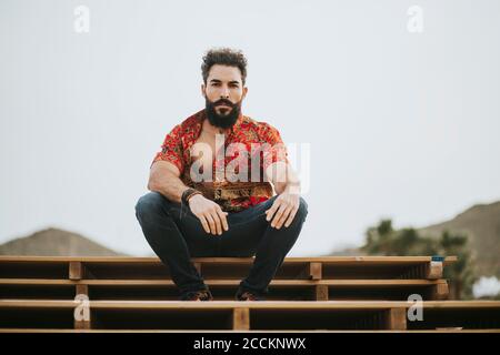 Beau barbu avec chemise colorée assise sur une pile de bois Banque D'Images