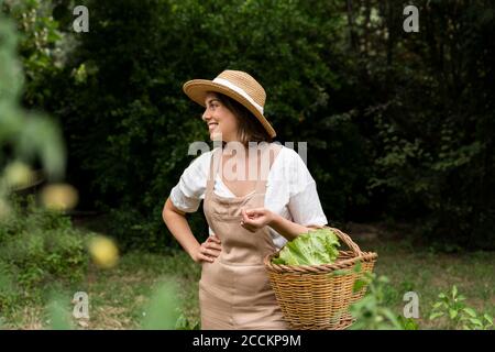 Jeune femme souriante portant un chapeau avec un panier en osier debout potager Banque D'Images