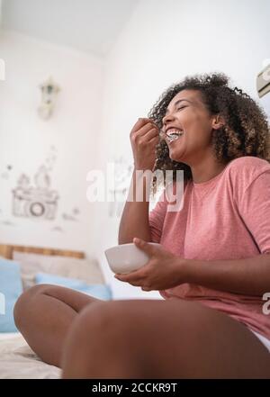 Jeune femme gaie mangeant de la nourriture tout en étant assise sur le lit à accueil Banque D'Images