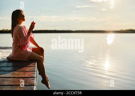 Jeune femme avec un verre de vin assis sur la jetée sur un lac au coucher du soleil Banque D'Images