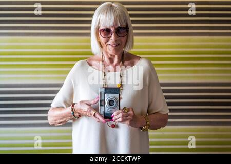Femme senior élégante portant des lunettes de soleil tenant l'appareil photo contre le mur à accueil Banque D'Images