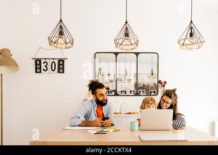 Homme avec un smartphone regardant la mère et la fille utilisant ordinateur portable sur la table à manger Banque D'Images