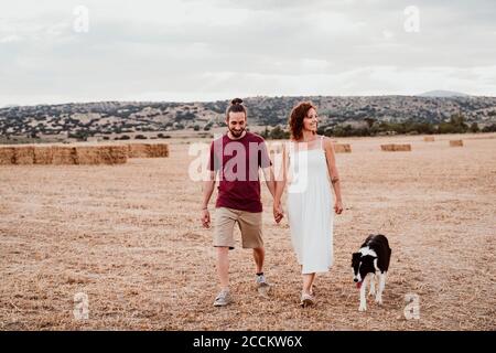 Couple souriant tenant les mains tout en marchant avec le chien dans le champ Banque D'Images